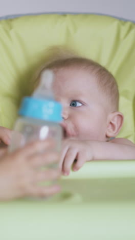 little blonde girl takes off bottle with water from junior brother sitting in green highchair in light room closeup