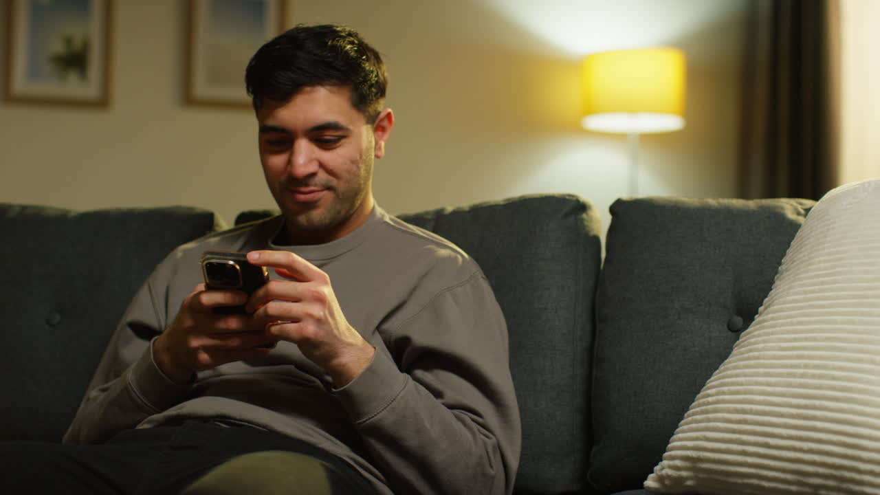 Young Man Spending Evening At Home Sitting On Sofa With Mobile Phone Scrolling Through Internet Or Social Media Streaming Content 2