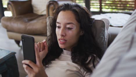 retrato de una mujer biracial pensativa acostada en el sofá y usando un teléfono inteligente