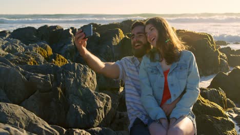 smiling happy young couple clicking selfie on rock at the beach 4k