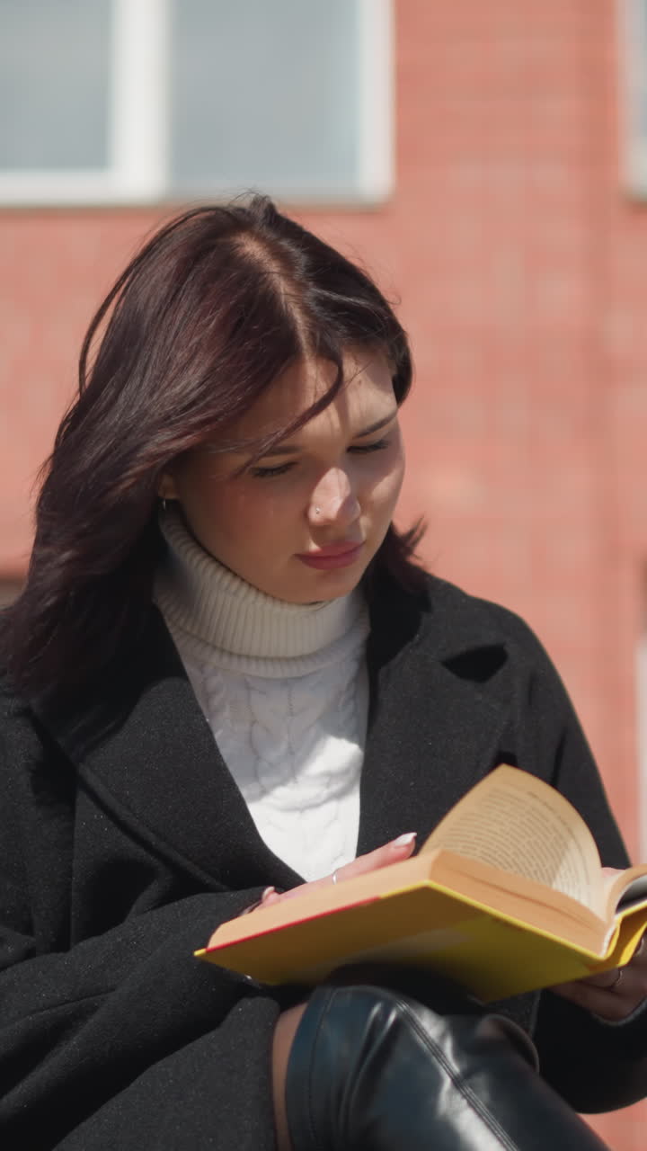 Woman in black coat sitting outside, thoughtfully reading her book as sunlight illuminates her face, her hair moves gently in the breeze, with a brick building in the background