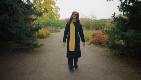 young lady with long wavy red hair wearing a yellow beret, scarf, and sunglasses, looking around happily with a blurred view of two people in the distant background