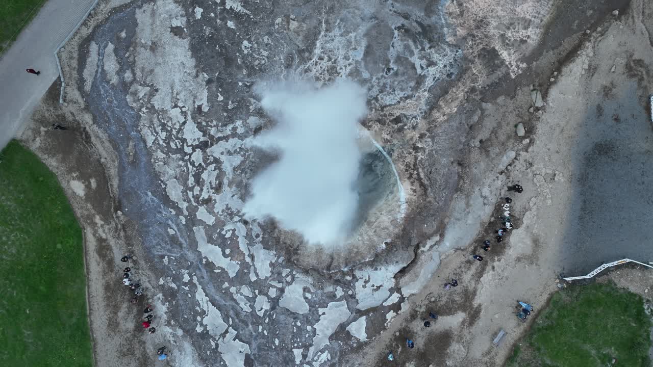 дронный вид гейзира, извергающегося в исландии, демонстрирующий мощный всплеск пара и воды, окруженный грубой геотермальной местностью.
