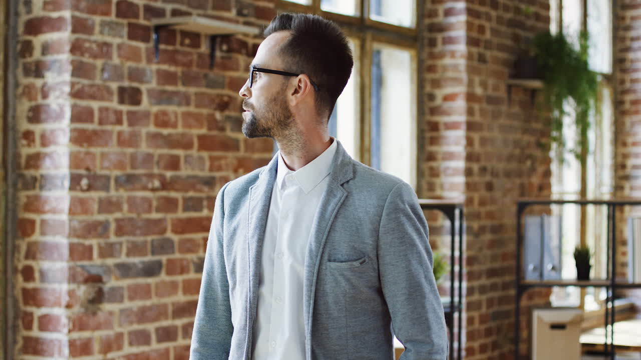 trabajador de oficina de buen aspecto con gafas mirando la ventana y luego girando la cabeza hacia la cámara en la habitación estilo loft