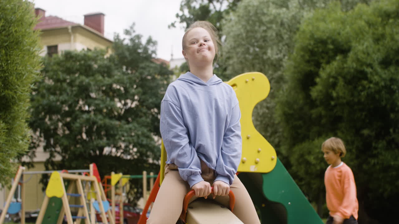 Nærbillede af en lille pige med Downs syndrom og hendes blonde veninde, der gynger på en trægynge i parken på en blæsende dag.