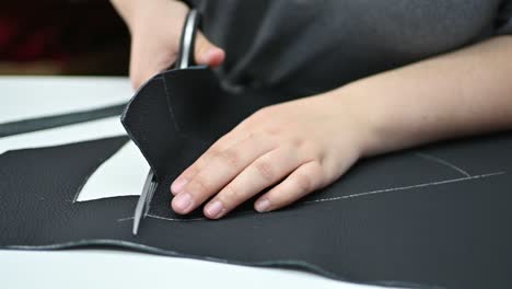 hands of an adult tailor cutting piece of black leather with scissors close up