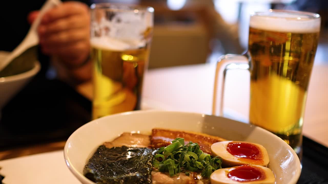 A detailed view of a ramen bowl with toppings and a glass of beer on a dining table.