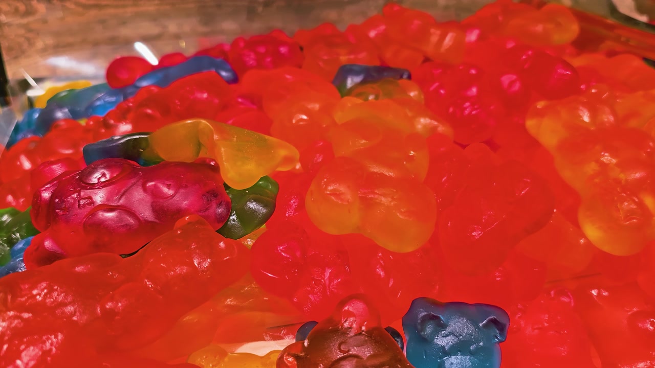 Nahaufnahme von bunten Gummibärchen, gestapelt in einem Süßigkeitenbehälter, mit durchscheinenden roten, orangefarbenen, gelben, grünen und blauen Süßigkeiten, glänzender Textur, warmem Licht und einer verspielten Snack-Auslage.