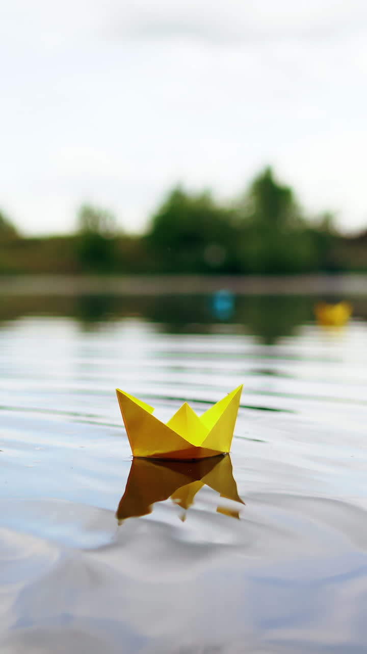Yellow paper boat is sailing on water surface under the evening sky. Origami ship made of yellow paper is swimming on the river at sunset. Vertical video