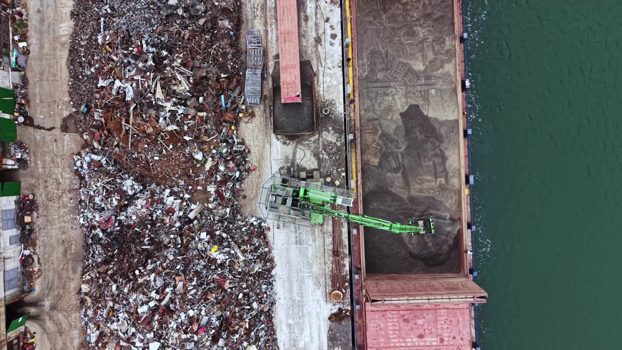 Operai smistano e gestiscono rottami metallici e materie prime in un impianto di riciclaggio sul lungomare sotto il sole pomeridiano.