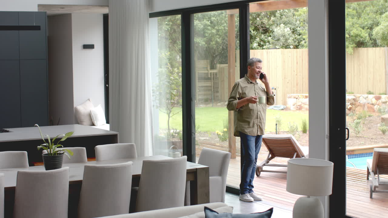 hombre bi-racial senior feliz en la terraza hablando en teléfono inteligente sosteniendo café, espacio de copia, cámara lenta