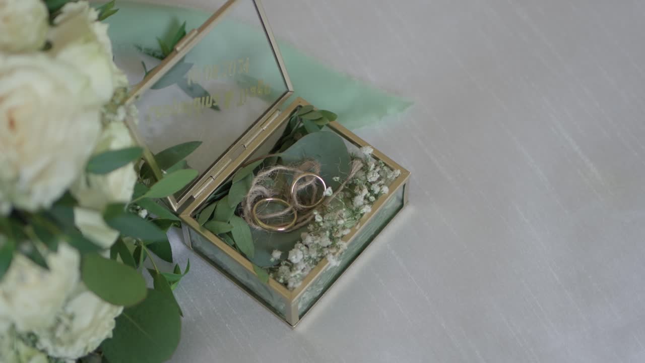 two gold wedding rings rest in a glass box adorned with baby's breath and eucalyptus leaves