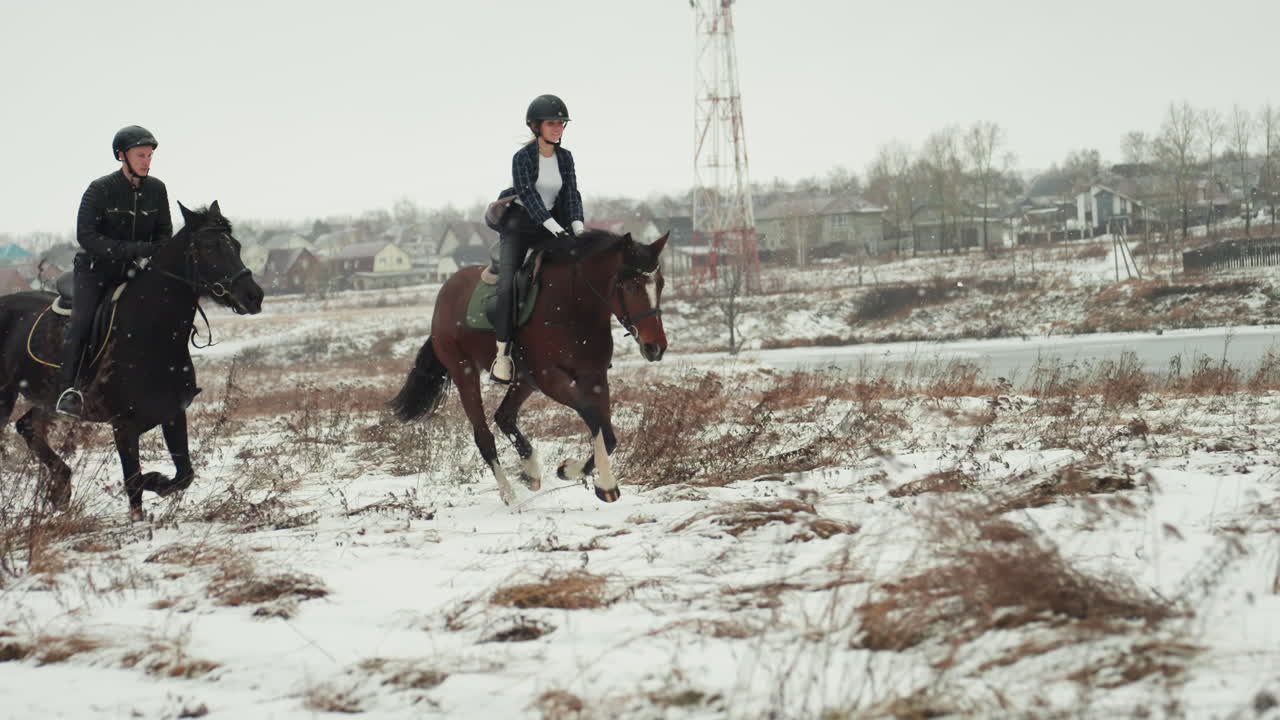 Due cavalieri caucasici a cavallo che praticano una lezione di equitazione in tandem in un campo innevato, istruttore che guida lo studente, caschi e selle, sfondo di villaggio rurale, lento galoppo e lavoro di squadra fiducioso.