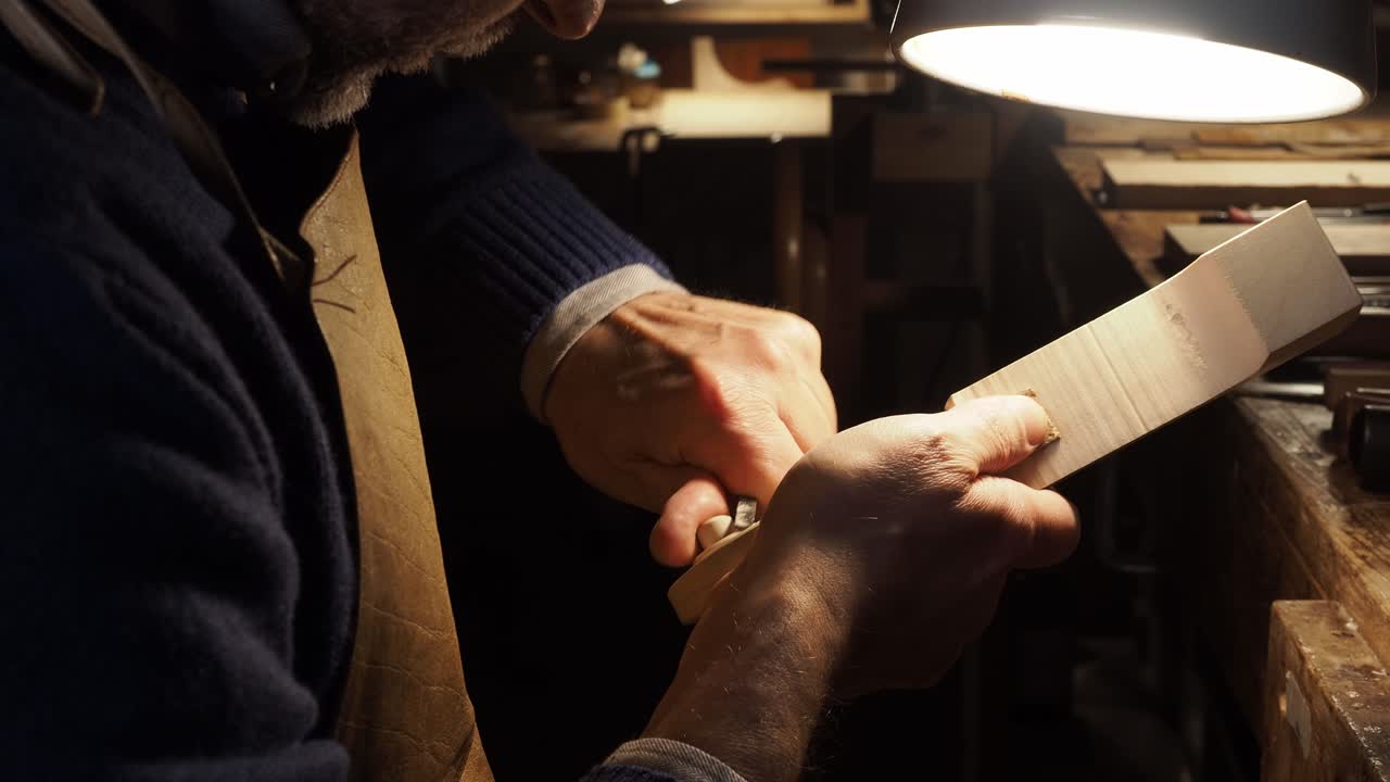 Close-up view of a luthier shaping the scroll of a violin by hand under warm light. The craftsman uses fine carving tools with patience and precision, revealing the artistry of violin making