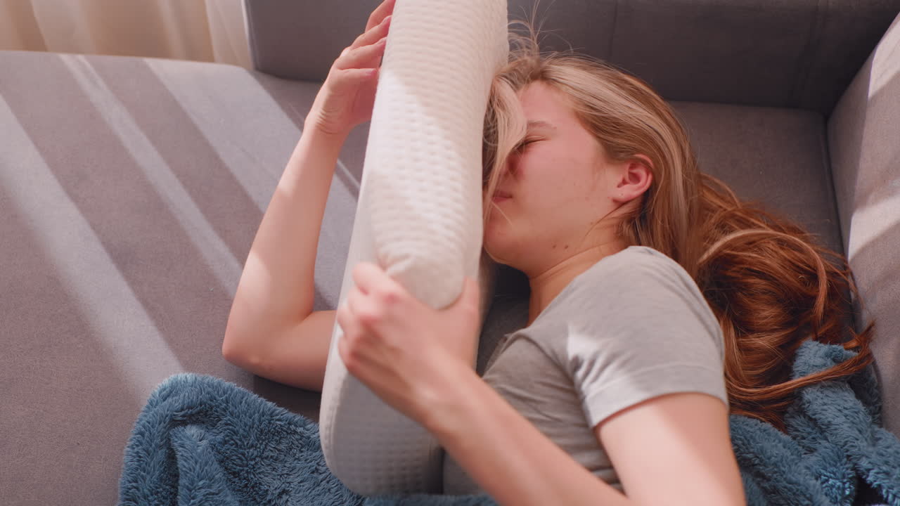 Top down view of college girl lying on couch hiding face in soft pillow unwilling to wake up from emotional exhaustion and stress, sunlight casting soft glow on her as blue blanket rests beneath