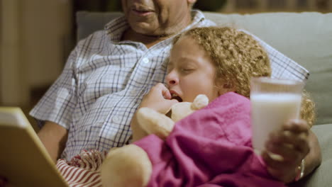el abuelo sostiene un vaso de leche y lee historias de buenas noches para su nieta que casi tiene sueño