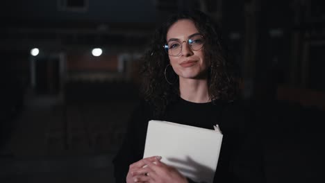 retrato de una chica morena segura con cabello rizado en gafas en un traje negro la actriz tiene un guión en sus manos y relata el texto de su personaje en el teatro durante un ensayo