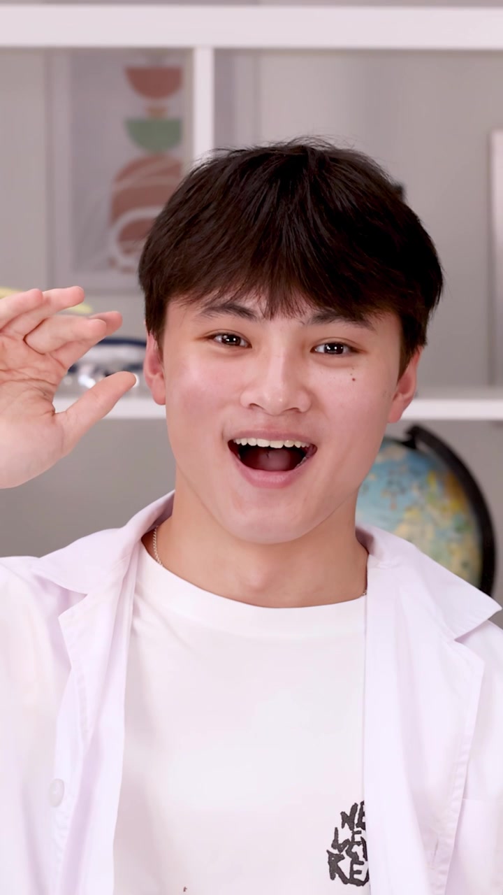 A happy teenage student wearing a white lab coat waves and smiles in a classroom.