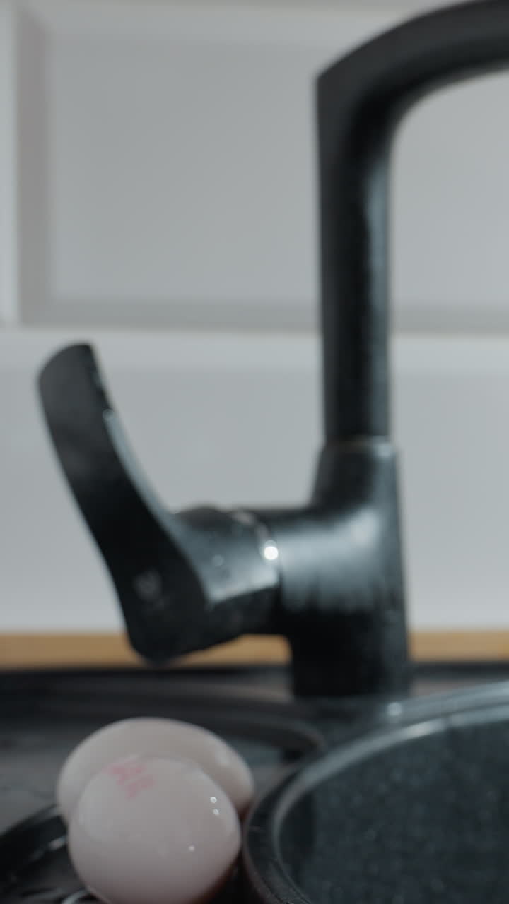 Primo piano delle mani di una donna che apre il rubinetto della cucina, prende un uovo e lo lava sotto l'acqua corrente. Uova fresche bianche appoggiate su un lavandino nero mentre scorre l'acqua