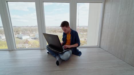 teen boy sitting on the floor by the window and playing on laptop. boy plays video games and celebrates victory. gaming at home.