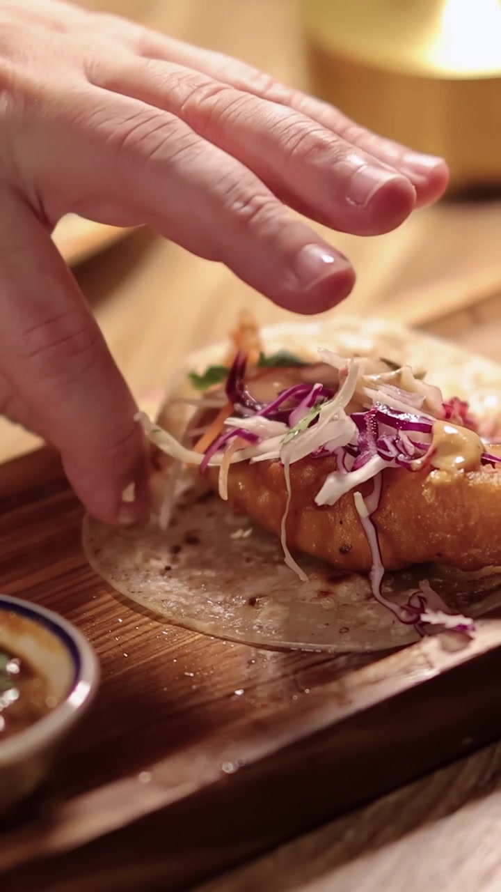 En hånd rækker ud for at tage en sprød fisketaco toppet med coleslaw og sauce på et træbræt, under varm, blød belysning med lav dybdeskarphed.