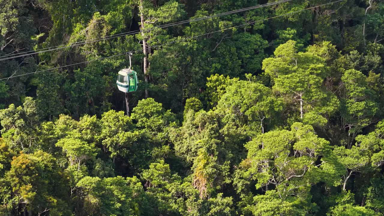 A green cable car travels smoothly over dense rainforest canopy in daylight, captured with a steady aerial shot highlighting vibrant foliage and natural landscape