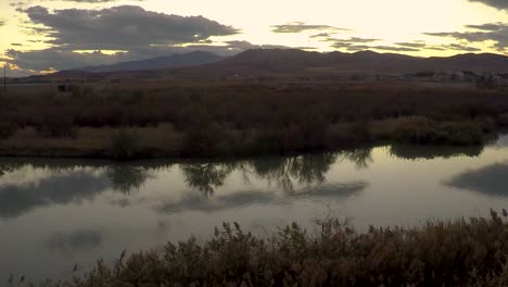 Lapso-De-Tiempo-Panorámico-De-Una-Puesta-De-Sol-Que-Se-Refleja-En-La-Superficie-Vidriosa-De-Un-Río-Con-Montañas-En-El-Fondo