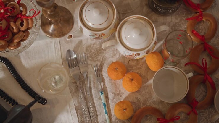 Overhead table with oranges and phone, scattered teacups, pretzels and ribbon, porcelain set and crumbs, coiled cord trailing off frame, hostess finishing celebration, melancholic reflective mood