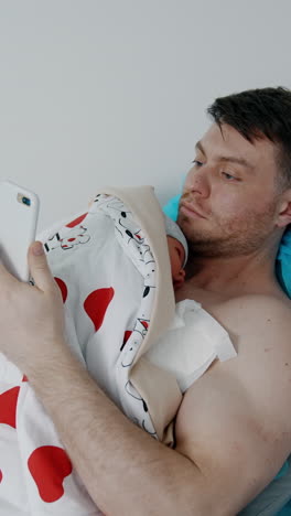 caucasian man holds his newborn child on the chest. father is looking at phone while baby is resting. vertical video
