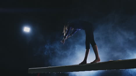 the girl is a professional athlete performs gymnastic acrobatic trick on a beam in backlight and slow motion in sports gymnastic clothing. smoke and blue. jump and spin on the balance beam