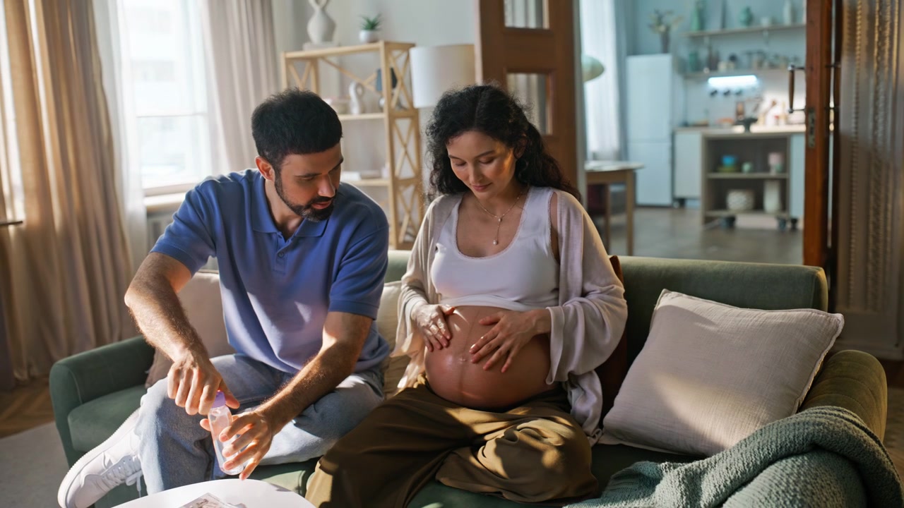 Expecting parents applying belly oil sofa. Husband caring pregnant wife at home.