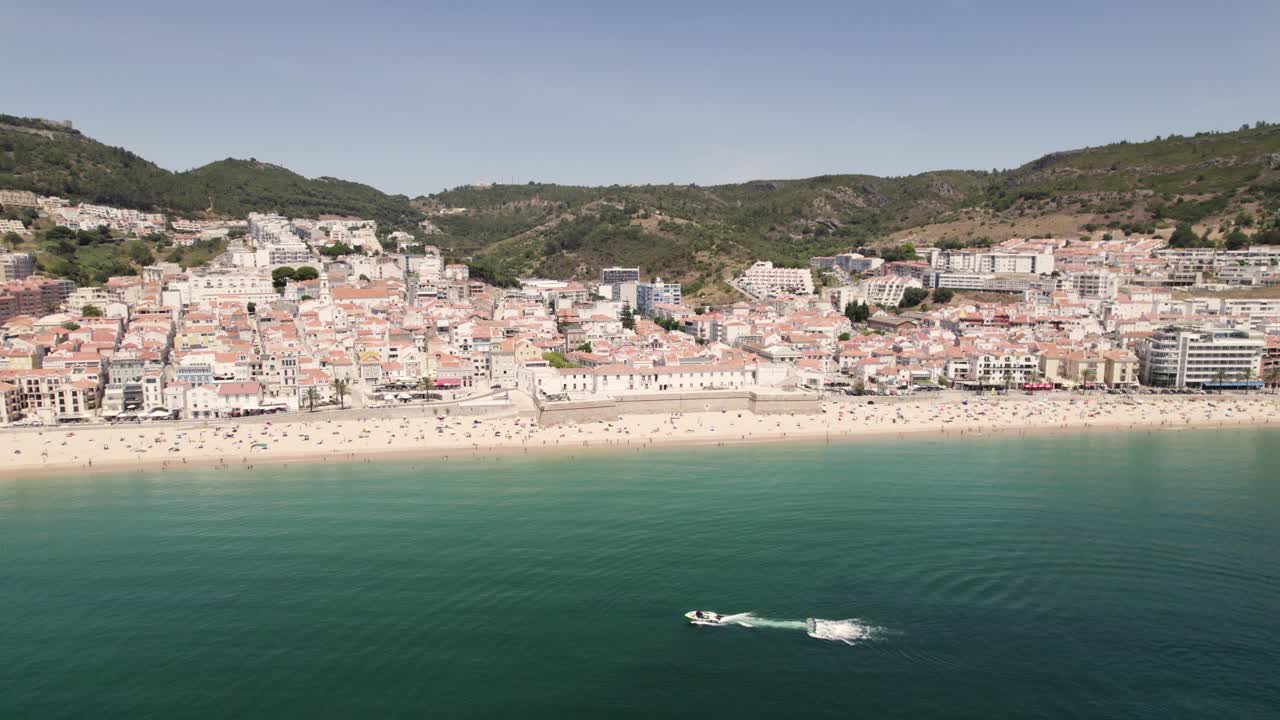 Persone che si godono la spiaggia di sabbia, le pittoresche case di Sesimbra sullo sfondo di colline e cielo blu