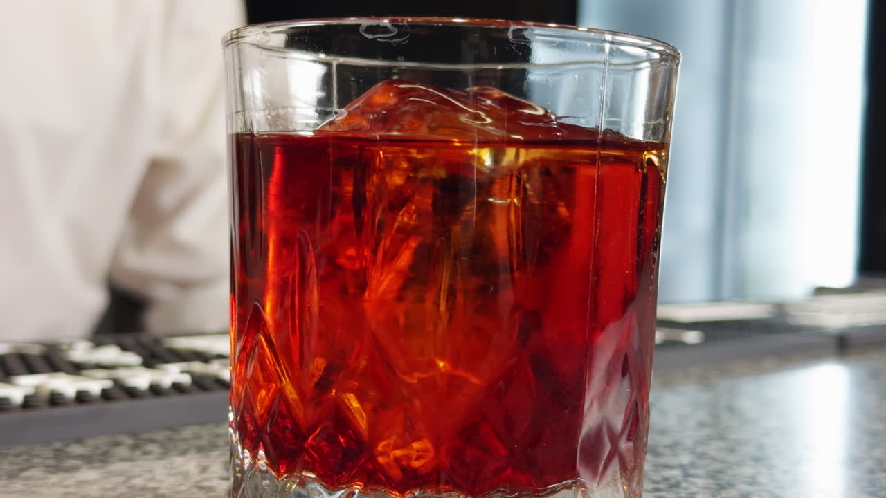 Old-fashioned glass filled with orange liquid. Unrecognized male bartender mixes the beverage with a spoon. Close up. Blurred backdrop.