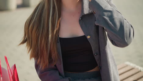 close-up of a woman wearing black crop top under grey shirt, casually touching her hair, a red shopping bag is visible beside her, and the background is slightly blurred