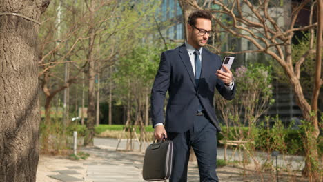 man in formal suit using phone while walking at city park