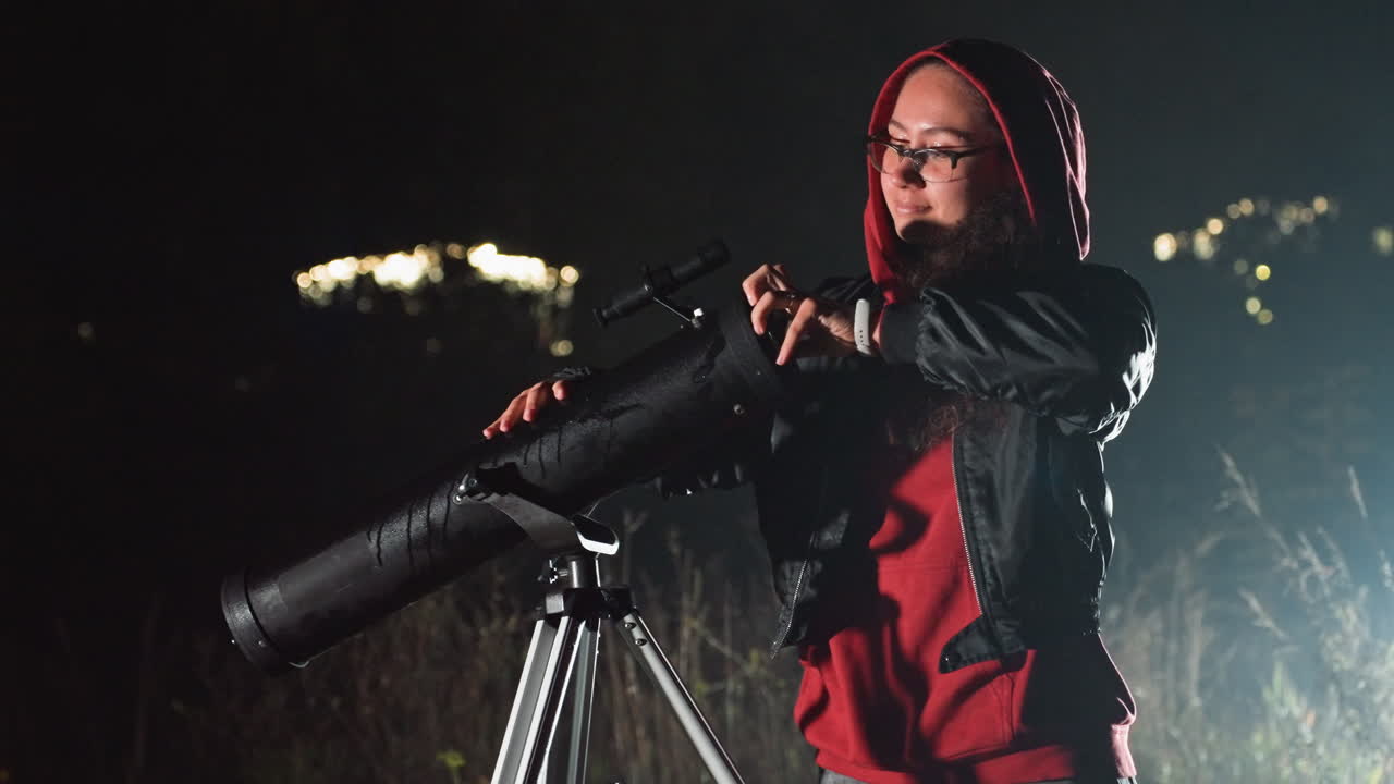 Donna caucasica che modifica il telescopio di notte, felpa rossa con cappuccio e giacca di pelle, occhiali, telescopio su treppiede puntato verso il cielo, campo silenzioso sotto le fioche luci della città, astronoma amatoriale concentrata che controlla le ottiche