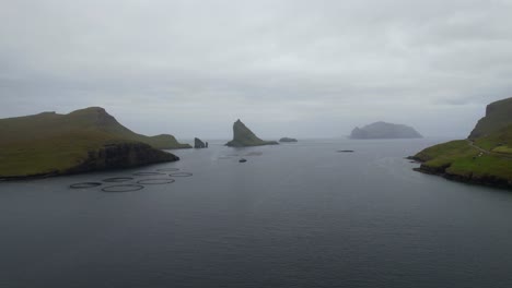 Drone-Volando-Hacia-Drangarnir-Y-Tindholmur-A-Través-De-Una-Granja-De-Salmón-En-Las-Islas-Feroe