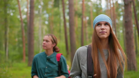 hikers walking along forest trail, woman in blue bandana and gray sweatshirt gazes around thoughtfully with a warm smile, while companion in green shirt adjusts her bag