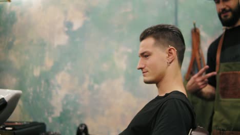 young bearded barber turning around the chair while the stylish client with piercing in his ear and tattoo is sitting there