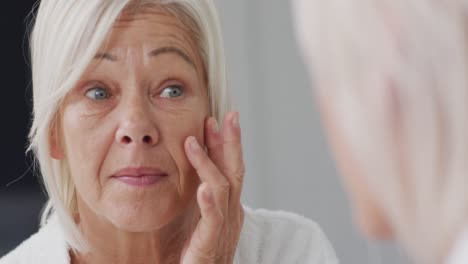 happy senior caucasian woman looking at mirror in bathroom and touching her face