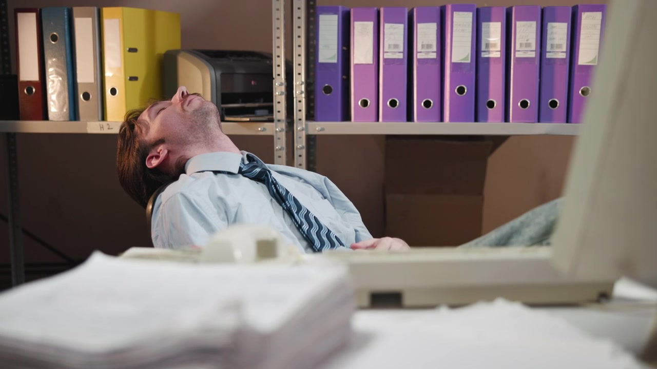 Un impiegato d'ufficio stanco si appoggia all'indietro sulla sedia all'interno di un ufficio archivio disordinato, circondato da raccoglitori, pile di carte, scaffali e una stampante sotto una luce soffusa
