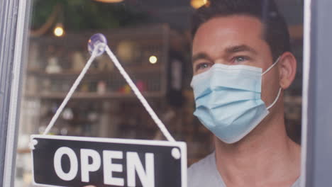 Male-Owner-Of-Small-Business-Wearing-Face-Mask-Turning-Round-Open-Sign-During-Health-Pandemic