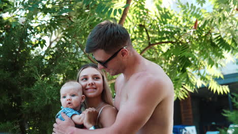 family of three on simmer vacation. topless man wearing glasses embraces his wife holding a child.