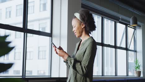 mujer de negocios afroamericana en la ventana usando un teléfono inteligente sola en la oficina