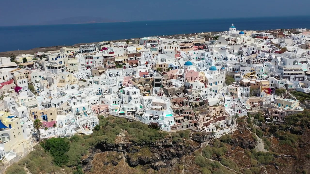 Bela tomada ampla de Oia, Santorini, apresentando casas brancas icônicas, igrejas de cúpulas azuis e dramáticas falésias vulcânicas vermelhas que descem em direção ao azul profundo do Mar Mediterrâneo em um dia claro.