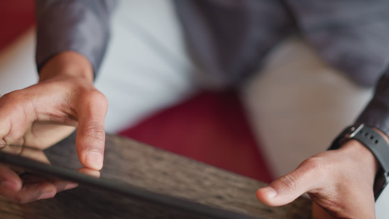 Uomo immigrato che tiene un tablet con entrambe le mani mentre è seduto al chiuso, concentrato sullo schermo con una presa salda, ripreso in primo piano che mostra i dettagli della mano, il coinvolgimento digitale e un ambiente moderno e informale.