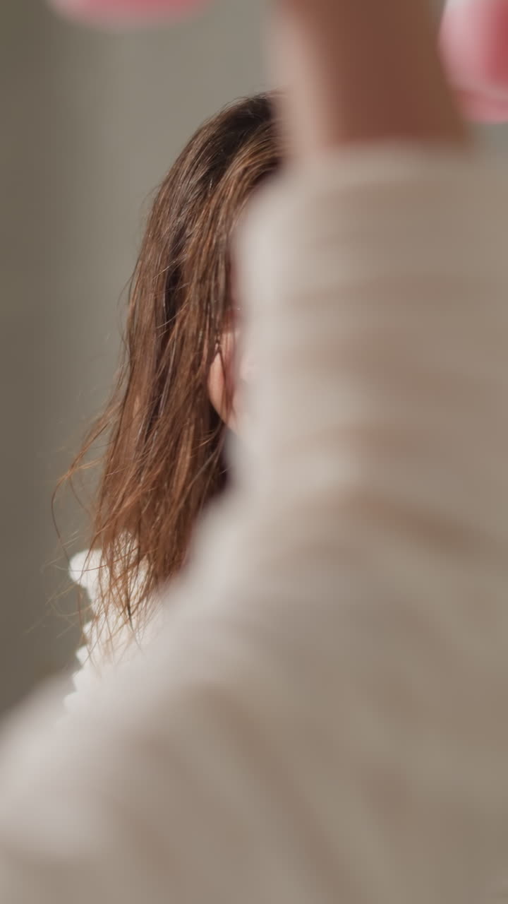 어두운 머리카락을 가진 여성은 넓은 욕실에서 에션을 적용합니다. 갈색 여성은 빛나는 액체를 스로우 모션으로 분사하여 실키 머리카락에 관심을 기울입니다.