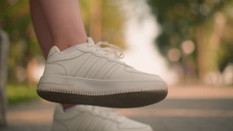 close-up of individual seated outdoors with one leg slightly lifted off ground wearing white sneakers, gentle movement of leg against blurred background illuminated softly by sunlight