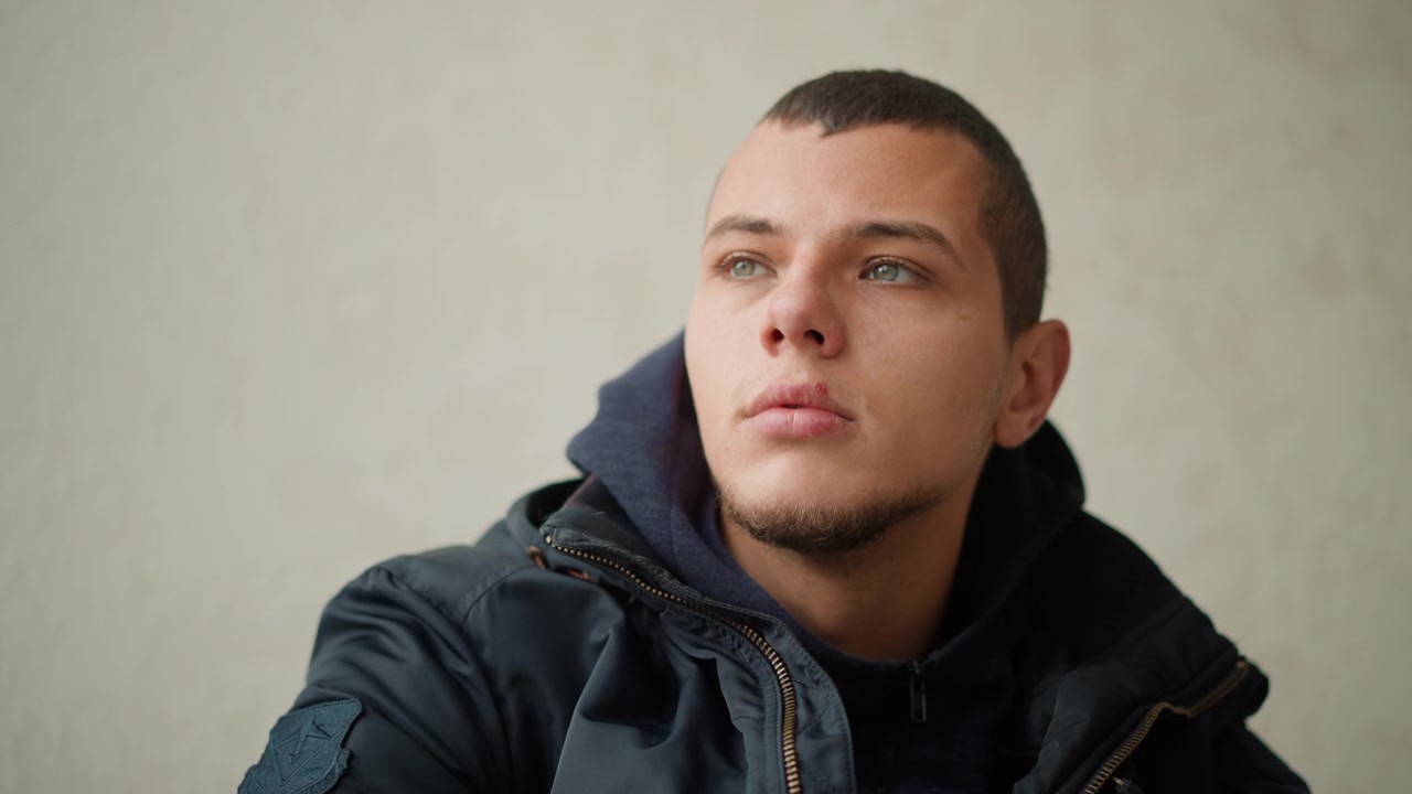 Primo piano di un giovane uomo che indossa una giacca scura e una felpa con cappuccio, seduto contro uno sfondo semplice. La sua espressione è calma con leggere variazioni nella direzione dello sguardo. L'illuminazione rimane morbida e uniforme.