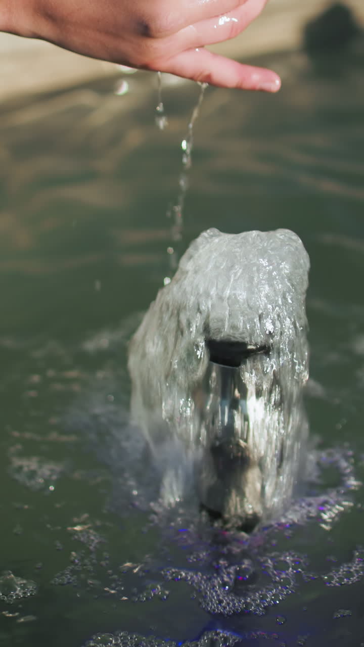 mano de un hombre con abrigo negro tocando el agua de manantial que fluye de una pequeña fuente, con una vista detallada de la superficie del agua burbujeante y los reflejos
